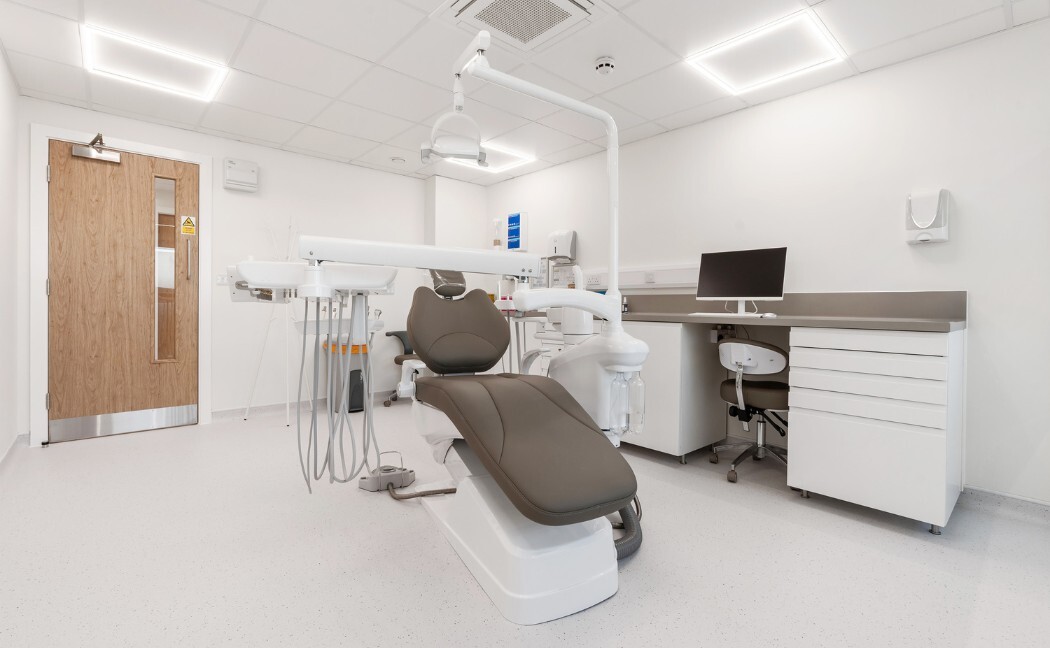 A bright white treatment room with a dental chair, overhead light and a clinical workstation with monitor.