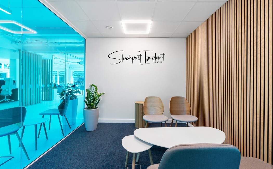 A quiet consultation corner with a timber-slat wall, the clinic's logo on a white wall, two chairs and a low white table beside blue-tinted glass.